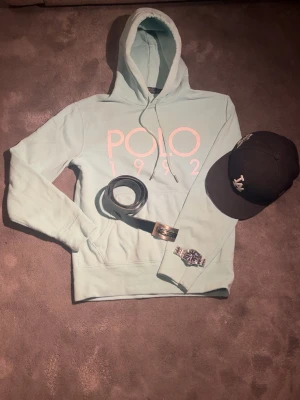 Ljusblå hoodie från Polo Ralph Lauren - Snygg ljusblå hoodie från Polo Ralph Lauren med stor vit logga och texten 'POLO 1992' på bröstet. Klassisk huva med dragsko och mjukt material som känns riktigt skönt mot huden. Perfekt för dig som vill ha en stilren och bekväm hoodie med ikonisk design.