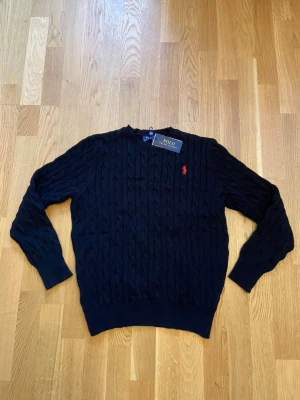 Svart kabelstickad Ralph Lauren tröja - Kabelstickad svart tröja från Polo Ralph Lauren med röd broderad logga på bröstet. Helt nytt skick och storlek S.