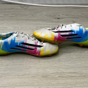 Adidas färgglada fotbollsskor - Säljer ett par Adidas fotbollsskor med en riktigt färgstark design i vitt, blått, gult och rosa. Skorna har klassiska svarta Adidas-ränder på sidan och är tillverkade i syntetmaterial med snörning och dobbar för spel på gräs.