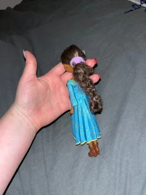 Docka - Säljer en docka med långt, lockigt brunt hår uppsatt med lila hårband. Dockan har en blå klänning med gula detaljer och bruna stövlar. Klänningen har trekvartsärm och gul kant längst ner. Perfekt för samling eller lek.