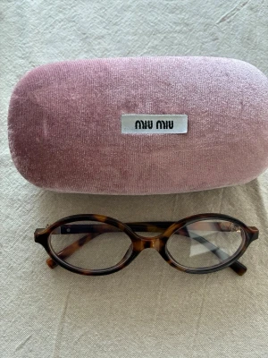 Bruna ovala solglasögon från Miu Miu - Säljer ett par snygga ovala solglasögon från Miu Miu med sköldpaddsmönstrad båge i bruna toner. Glasögonen har gulddetaljer vid skalmarna och kommer med ett rosa sammetsfodral. Blåljus och ingen styrka
