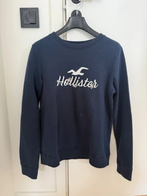 Mörkblå sweatshirt från Hollister - Mörkblå sweatshirt från Hollister med broderad logga och text på bröstet. Tröjan har rund halsringning, långa ärmar och ribbade muddar vid ärmslut och nederkant. Perfekt för en avslappnad och stilren look.