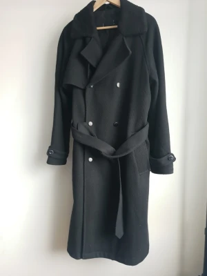 Svart trenchcoat med bälte Asos - Stilren svart trenchcoat med dubbelknäppning och avtagbart bälte i midjan. Jackan har bred krage med mjukt tyg, två sidofickor och knappar vid ärmsluten. Tillverkad i ett tjockare material som passar perfekt för kyligare dagar.