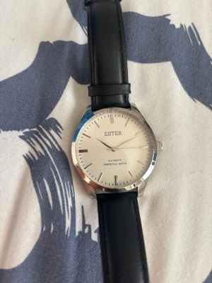 ESTER silverklocka med svart läderband - Stilren klocka från ESTER med silverfärgat ur och svart läderband. Urtavlan är vit med minimalistiska markeringar och texten 'Automatic Perpetual Aston'. Perfekt accessoar för dig som gillar klassisk och enkel design.