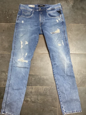 Replay jeans - Säljer ett par blå jeans från Replay med slitningar och distressed detaljer framtill. Klassisk femficksmodell med raka ben och normal midja. Jeansen har vita kontrastsömmar och Replay-lapp bak i midjan. Perfekta för en avslappnad och trendig look.