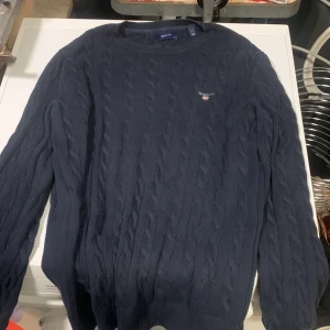 Mörkblå kabelstickad tröja Gant - Snygg mörkblå kabelstickad tröja från Gant med rund halsringning och diskret logga broderad på bröstet. Tröjan har långa ärmar och ribbade muddar vid ärmslut och nederkant. Perfekt för lager på lager och klassisk stil.