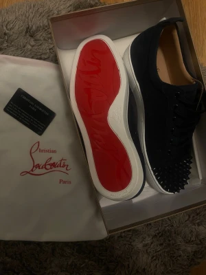 Christian Louboutin sneakers - Säljer ett par mörk blåa sneakers från Christian Louboutin med ikonisk röd sula och svarta nitar på tån. Skorna har lågt skaft, snörning och är tillverkade i mocka. Kommer med originalpåse och certifikat.