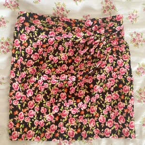 Blommig kjol från Zara -  Färgglad blommig kjol från Zara med rosa och gröna blommor på en svart bakgrund. Kjolen har en omlott-design som ger en fin detalj framtill.