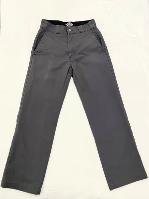 Mörkgrå byxor från Dickies - Snygga mörkgrå chinos från Dickies Skateboarding-kollektion. Byxorna har raka ben, klassiska sidfickor och en knappgylf. Liten slitning på höger framficka. 
