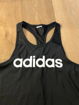 Svart träningslinne från Adidas - Snyggt svart träningslinne från Adidas med stort vitt logotryck på bröstet. Linnet har brottarrygg och meshpaneler upptill för extra ventilation. Perfekt för gymmet eller löprundan, tillverkat i ett lätt och snabbtorkande material.