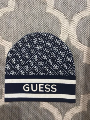 Mörkblå mönstrad mössa från Guess - Snygg mörkblå mössa från Guess med vitt logomönster och breda vita ränder längst ner. Använd ca 2 gånger 