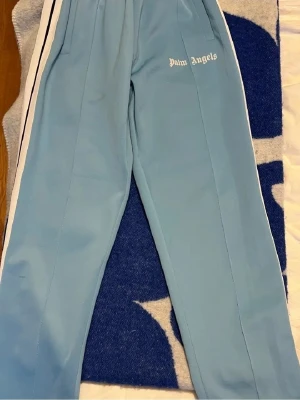 Ljusblå mjukisbyxor Palm Angels - Säljer ett par ljusblå mjukisbyxor från Palm Angels med vita sidoränder och logga tryckt på vänster sida. Byxorna har resår i midjan och är gjorda i ett glansigt, mjukt material som känns riktigt skönt. Perfekta för en chill och trendig look.