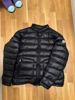 Svart Moncler dunjacka herr - Säljer en svart Moncler dunjacka med klassisk quiltad design och hög krage. Jackan har dragkedja framtill, två sidofickor med dragkedja och Moncler-logga på ärmen. Perfekt för kyliga dagar och riktigt snygg till streetwear. Dragskedja är sönder men den fungerar att stänga som normalt. 