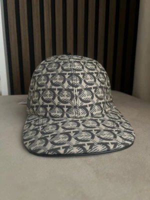 Beige och svart keps från Ferragamo  - Snygg keps från Ferragamo  i beige och svart med ett grafiska mönster över hela kepsen. Klassisk form med böjd skärm och justerbart band bak. Tillverkad i ett slitstarkt tyg som ger en exklusiv känsla.