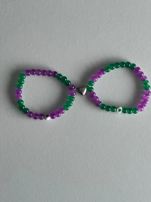 Lila och grönt pärlarmband med magnet-hjärt - Två armband med glansiga pärlor i lila och grönt, sammanlänkade med ett silverfärgat hjärtlås. Varje armband har en liten vit och silverfärgad pärldetalj som accent. Perfekt för dig som gillar färgglada accessoarer och unika detaljer.