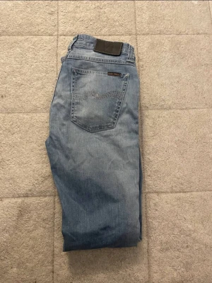 Ljusblå jeans från nudie  - Säljer ett par ljusblå jeans från nudie med klassisk femficksmodell och diskret logga på bakfickan. Jeansen har raka ben och är tillverkade i mjukt denimtyg. Perfekta för en avslappnad och trendig look.