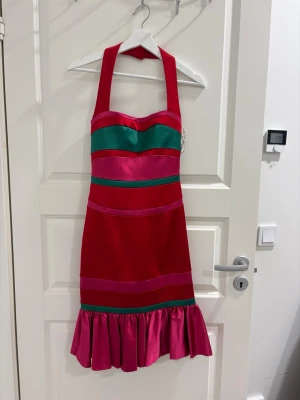 Röd och rosa halterneckklänning - Färgstark kortklänning med halterneck och breda axelband. Klänningen har röda, rosa och gröna blockränder samt en volangkant nertill. Tillverkad i glansigt material som ger en lyxig känsla. Perfekt för dig som vill sticka ut med en unik och trendig look.