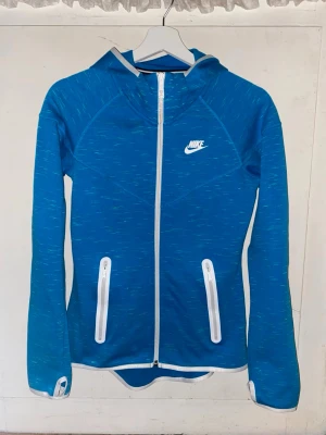 Blå zip hoodie från Nike - Sportig blå hoodie från Nike med hel dragkedja och vita detaljer. Tröjan har två fickor med dragkedja, tumhål i ärmsluten och en smal passform. Perfekt för träning eller chill. Tillverkad i ett stretchigt funktionsmaterial. Storlek S