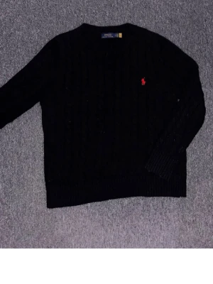 Svart kabelstickad tröja Polo Ralph Lauren - Kabelstickad svart tröja från Polo Ralph Lauren med rund halsringning och röd broderad logga på bröstet. Tröjan har ribbade muddar vid ärmslut och nederkant. Perfekt för dig som gillar klassisk och stilren design.