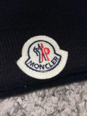 Svart Moncler mössa - Säljer en svart ribbstickad mössa från Moncler med den klassiska broderade logotypen framtill. Mössan har uppvikt kant och är tillverkad i mjukt material som håller dig varm. Perfekt accessoar för kyliga dagar och snygg till streetwear-stilen.