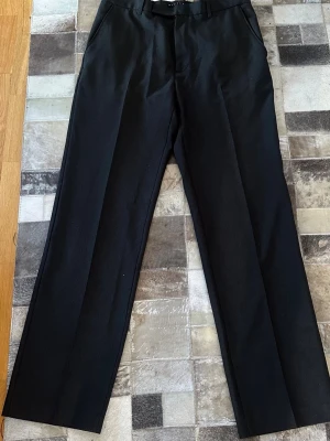 Svarta klassiska kostymbyxor - Snygga svarta kostymbyxor med raka ben och pressveck. Byxorna har två sidofickor fram och två bakfickor med knapp. Perfekta för en stilren look och passar till skjorta eller t-shirt.