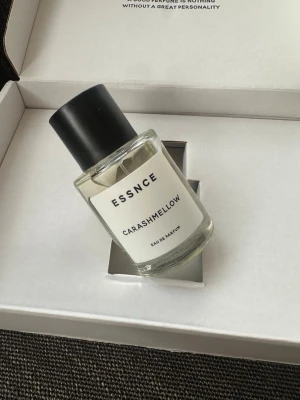 ESSNCE Carashmellow Eau de Parfum - Minimalistisk parfymflaska från ESSNCE med doften Carashmellow. Flaskan är cylindrisk i genomskinligt glas med svart rund kork och vit etikett med svart text. Modern och stilren design som passar perfekt i badrumshyllan.