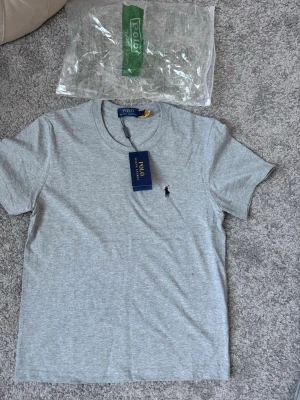 Grå t-shirt från Polo Ralph Lauren - Snygg grå t-shirt från Polo Ralph Lauren med klassisk passform och korta ärmar. Tillverkad i mjuk bomull och har den ikoniska lilla broderade loggan på bröstet. Perfekt basplagg för alla tillfällen.