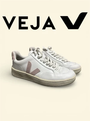 Vita Veja sneakers med rosa detaljer - Stilrena vita sneakers från Veja med ljusrosa V-logga på sidan och matchande rosa detalj vid hälen. Skorna har klassisk låg profil, vita snören och sula i beige ton. Ovandelen är i slätt läder och insidan är fodrad med textil för extra komfort.