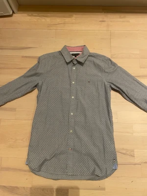 Grå skjorta Tommy Hilfiger - Snygg grå skjorta från Tommy Hilfiger med små prickar över hela tyget. Klassisk krage, knäppning framtill och långärmad modell. Diskret logga på bröstet och kontrastfärgad insida vid kragen. Perfekt för en stilren look.