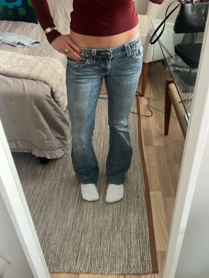 Blingade Miss Me jeans med låg midja - Säljer ett par blå Miss Me jeans med låg midja och raka ben. Jeansen har snygga slitningar och är dekorerade med nitar och strass på fickorna både fram och bak. Perfekta för dig som gillar detaljer och vill sticka ut lite extra. BYXORNA ÄR OMSYDDA I LÄNGDEN. Skriv för fler bilder❤️