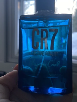 CR7 parfym blå flaska cool  - Snygg parfym från Cristiano Ronaldo, CR7 Play It Cool, i en genomskinlig blå glasflaska med svart lock och silverdetaljer. Flaskan har en modern, fyrkantig form och en ikonisk logga på locket. Perfekt för dig som gillar fräscha och sportiga dofter.