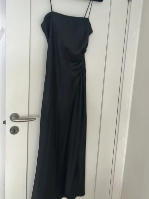 Svart långklänning från Zara - Elegant svart långklänning från Zara med tunna axelband och lätt rynking vid sidan. Den här även en slits. Klänningen har en slät, stilren design och är tillverkad i ett glansigt material som ger en lyxig känsla. Perfekt för dig som gillar minimalistisk och trendig stil.