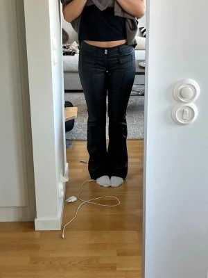 Svarta bootcut skinbyxor med låg midja - Säljer ett par svarta bootcut skinbyxor  med låg midja och två knappar framtill. Byxorna har en snygg, figurnära passform upptill och släpper ut vid benen. Materialet ser ut att vara stretchigt och bekvämt, perfekt för en trendig look.