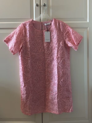 Rosa blommig kortklänning från IVIVI - Supersöt rosa kortklänning från IVIVI med 3D-blommönster och paljettdetaljer. Klänningen har rund halsringning och korta ärmar. Helt ny och etiketten sitter kvar. Storlek M.