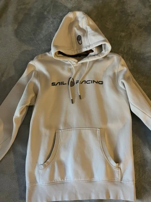 Beige hoodie från Sail Racing - Snygg beige hoodie från Sail Racing med svart logga tryckt på bröstet och en broderad detalj på huvan. Hoodien har dragsko i huvan och en stor känguruficka framtill. Tillverkad i mjukt bomullsmaterial, perfekt för en chill och avslappnad stil.