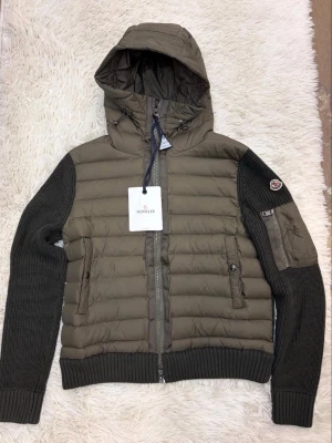 Moncler grön hybrid cardiganjacka - Stilren cardiganjacka från Moncler i olivgrönt med vadderad front och huva. Ärmarna är stickade i mörkgrönt och har en patchficka med dragkedja samt Moncler-logga på armen. Jackan har ribbade muddar och två fickor fram med dragkedja.