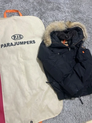 Svart Parajumpers jacka  - Säljer en svart parkas från Parajumpers med avtagbar pälskant på huvan och orange detaljer. Jackan har flera fickor, patch på ärmen och varm, mjuk foder. Perfekt för kalla dagar och riktigt snygg streetstil.