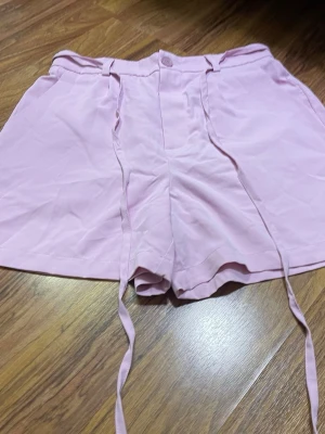 Ljusrosa shorts med snörning - Säljer ett par ljusrosa shorts med hög midja och snygg snörning framtill. Shortsen har bälteshällor och stängs med knapp och dragkedja. Perfekta för varma sommardagar och har en loose fit som ger en avslappnad vibe.