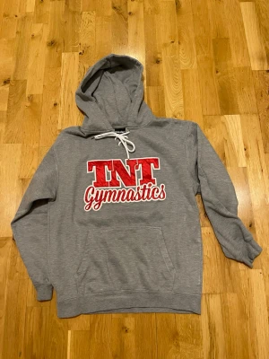 Grå hoodie TNT Gymnastics stl S  - Snygg grå hoodie med stor röd och vit TNT Gymnastics-logga på bröstet. Klassisk känguruficka framtill och vit dragsko i huvan. Perfekt för dig som gillar gymnastik och vill ha en bekväm och stilren tröja.