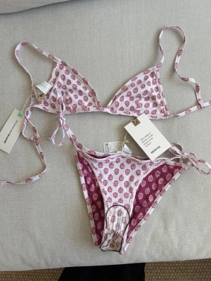 Rosa och vit blommig bikini från Brownie - Supersöt bikini från Brownie med blommigt mönster i rosa och vitt. Triangeltopp och trosa med knyt i sidorna, båda delarna är vändbara med olika färgkombinationer. Perfekt för strandhäng och sommar vibes!