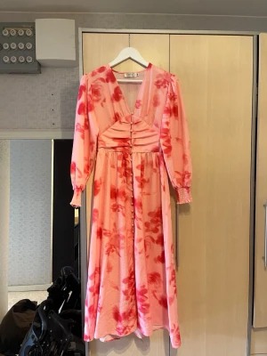 Rosa och röd långklänning med v-ringning - Snygg långklänning i rosa med röda blommiga mönster och v-ringad hals. Klänningen har långa ärmar med smock vid ärmslut och markerad midja med rynkade detaljer framtill. Det är även en slits framtill. Perfekt för dig som vill sticka ut med färg och mönster. ❤️🩷