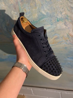 Christian louboutin skor - Snygga mörkblå sneakers från Christian Louboutin i mocka med svarta nitar på tån och klassisk röd sula. Skorna har lågt skaft, matchande mörkblå snörning och vita kanter på sulan. Perfekta för dig som vill sticka ut med en lyxig och edgy stil.