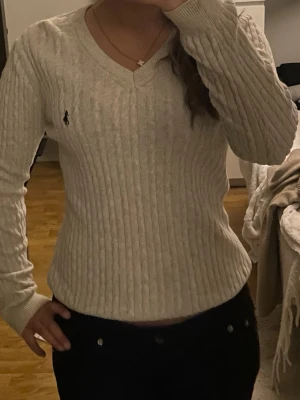 Beige kabelstickad tröja från Ralph Lauren - Kabelstickad tröja i beige från Ralph Lauren med V-ringning och lång ärm. Klassisk broderad logga på bröstet. Tröjan har en figurnära passform och ribbade muddar vid ärmslut och nederkant. Perfekt för en stilren och tidlös look.