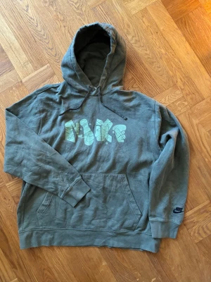 Vintage Grön khaki Nike hoodie street Y2K  - Snygg grön hoodie från Nike med stor grafisk logga i ljusgrönt och vitt på bröstet. Klassisk känguruficka framtill, huva med dragsko och ribbade muddar. Perfekt för en avslappnad streetwear-look.