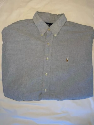 Ralph Lauren | Skjorta | L - Klassisk ljusblå skjorta från Ralph Lauren med button-down krage och broderad logga på bröstet. Skjortan har långa ärmar, normal passform och är tillverkad i mjuk bomull. Perfekt för en clean och stilren look. Mått: 75 Längd, 63 Bredd