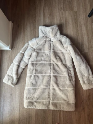 Ljusbeige lång pälsjacka - Säljer en supermjuk och fluffig ljusbeige pälsjacka i fuskpäls. Jackan har hög krage, raka ärmar och går ner över höfterna för extra värme. Perfekt för kalla dagar när du vill känna dig både stylish och cozy.