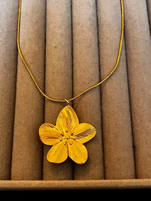 Guldigt halsband med blomma - Unikt halsband med en stor blomformad berlock i guldton. Kedjan är tunn och elegant, och blomman har fina detaljerade linjer som ger ett handgjort intryck. Perfekt för dig som vill sticka ut med ett smycke som känns både trendigt och personligt.