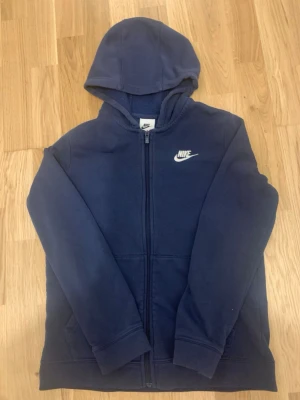 Mörkblå Nike hoodie med dragkedja - Snygg mörkblå hoodie från Nike med dragkedja framtill och klassisk vit Nike-logga på bröstet. Tröjan har huva och två fickor framtill. Perfekt för en avslappnad och sportig stil. Mjuk och skön bomullsblandning. Passar dig som är 154 ish bäst. Kan bara mötas, förlåt.