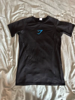 Svart träningströja från Gymshark - Snygg svart träningströja från Gymshark med blå logga på bröstet. T-shirten har korta ärmar och är tillverkad i ett stretchigt funktionsmaterial som andas, perfekt för gymmet eller löpning.