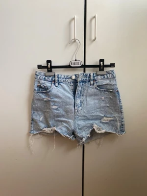 Ljusblåa denimshorts med slitningar - Säljer ett par ljusblåa denimshorts från Stradivarius med hög midja och råa, fransiga kanter. Shortsen har slitna detaljer framtill och klassiska fem fickor. Perfekta för sommaren och ger en avslappnad vibe.